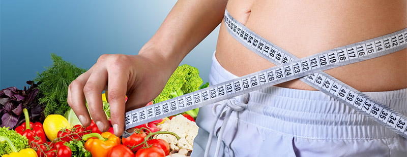 Ein Bild mit einer Person die ein Maßband um Ihren Bauch hat und im Hintergrund sieht man Gemüse und Obst