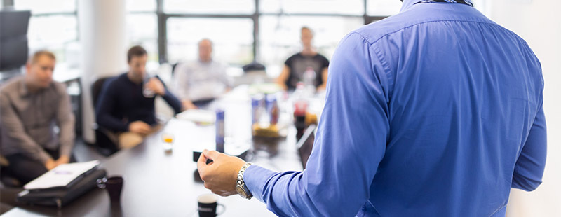 Ein Bild auf dem ein Mann in einem blauen Hemd vielen Menschen die an einem tisch sitzen im Hintergrund etwas erklärt