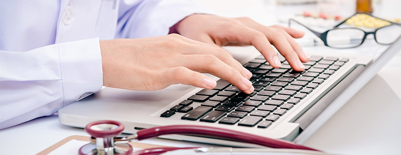 Ein Bild auf dem eine Person auf der Tastatur etwas tippt neben dran liegt ein Stethoskop und eine Schwarze Brille