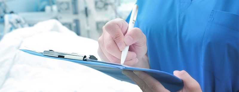 Ein Bild auf dem eine Person ein Brett mit einem Zettel in der Hand hält und etwas notiert, im Hintergrund steht ein Krankenbett