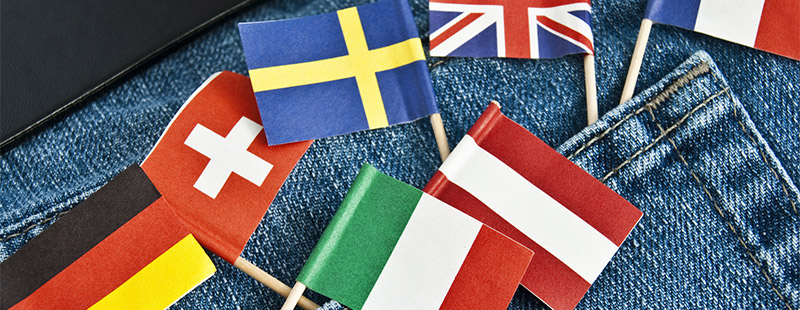 Ein Bild auf dem Man einen Jeans Stoff sieht, in der Hosentasche stecken Bunte Länder Fahnen wie die Schweiz, Italien....