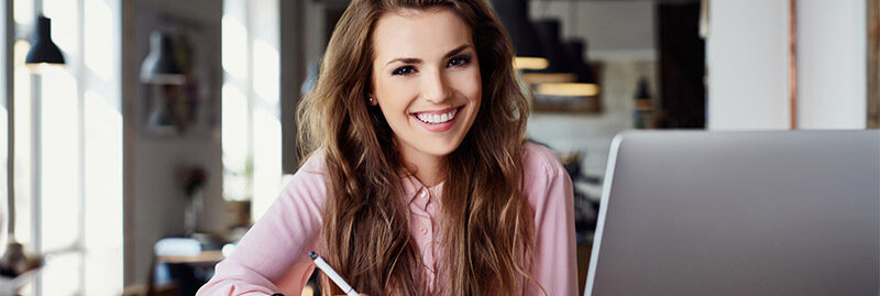 Eine lächelnde Frau die am Schreibtisch sitzt und einen Kugelschreiber in der Hand hält. Ein Laptop ist auch zu sehen