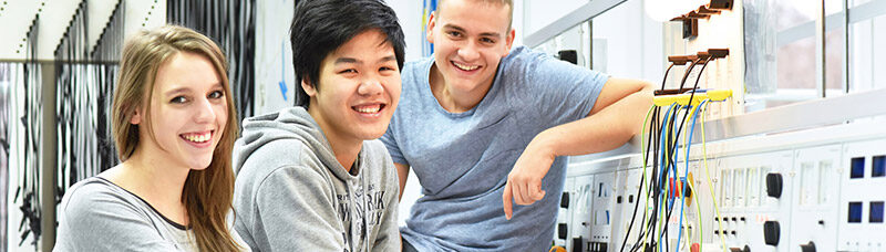 Ein Bild wo 3 Personen an einer Maschine sitzen und lächeln, im Hintergrund sieht man weitere Maschinen und die dazugehörigen Arbeitsmaterialien