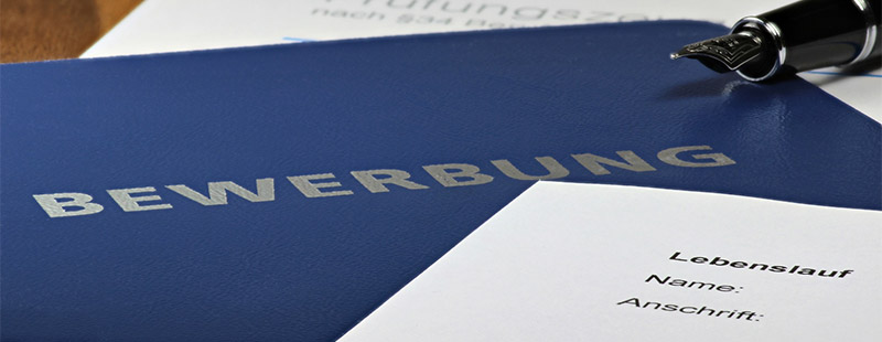 Hier sieht man eine Blaue Bewerbungsmappe darauf liegt halb ein Lebenslauf und ein Füller