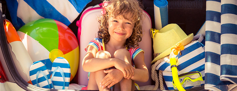Ein Kind das zwischen ganz vielen Bunten Urlaubsachen sitzt, z.B: einem Ball, einem gelben Hut, Flippflopps