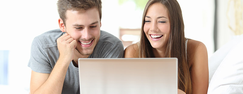 2 Personen die auf dem Bett liegen und lachend in einen Laptop schauen