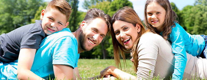 Eine Familie die im Grünen gras liegt mit den Kindern auf dem Rücken. Im Hintergrund sind Bäume