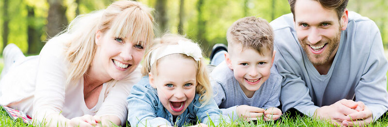 Eine lachende Familie liegt im Gras und im Hintergrund sind grüne Bäume