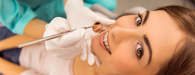 Eine Frau die eine Zahnspange hat und auf einem Zahnarztstuhl sitzt darüber Beugt sich eine Zahnarzthelferin mit einem Spiegel