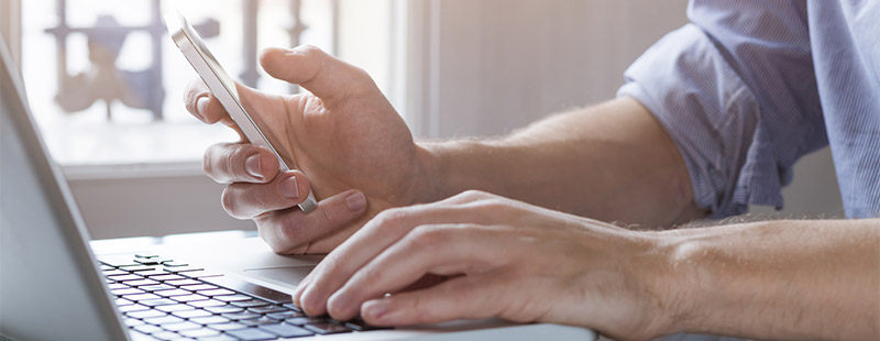 Auf diesem Bild sieht man ein Mann der am Laptop arbeitet und in der Linken Hand ein Smartphone hält