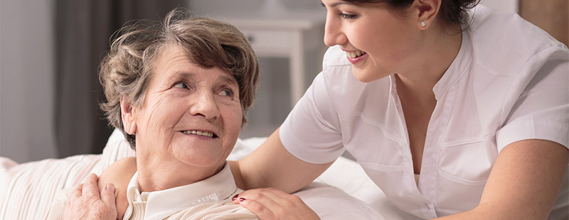 Ein Bild auf dem eine ältere Dame auf dem Sofa sitzt und eine Pflegerin Ihre Hand um der älteren Dame Ihre Schulter legt und lächelt