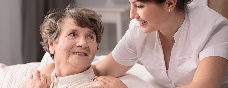 Ein Bild auf dem eine ältere Dame auf dem Sofa sitzt und eine Pflegerin Ihre Hand um der älteren Dame Ihre Schulter legt und lächelt