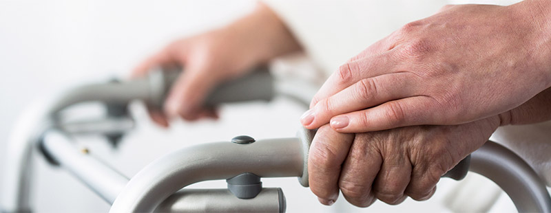 Ein Bild auf dem der obere Teil eines Rollators zu sehen ist an dem eine ältere Hand Festhält und eine andere Hand die ältere Hand hält.