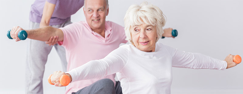 Ein Bild auf dem eine ältere Frau Ihre Arme ausgestreckt und jeweils eine Hantel in dem Armen hält, hinter Ihr sitzt ein Mann der ebenso mit gestreckten Armen Hanteln in den Händen hält. Bei dem Mann wird als Unterstützung eine Person helfen