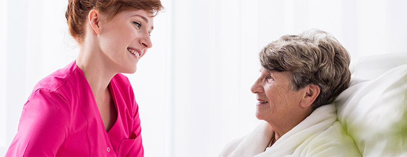 Eine ältere Dame die im Krankenbett sitzt und eine gegenüber sitzende Pflegekraft die lächelt