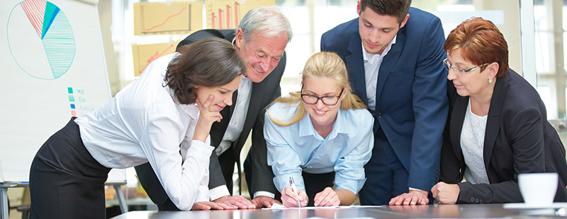 Ein Bild auf dem mehrere Personen auf einen Zettel am Tisch schauen und im Hintergrund sieht man ein Wake Board und verschiedene Zeichnungen