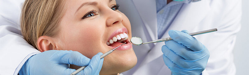 Ein Bild auf dem eine Frau beim Zahnarzt ist und dahinter der Arzt die Zähne mit einem Spiegel kontrolliert