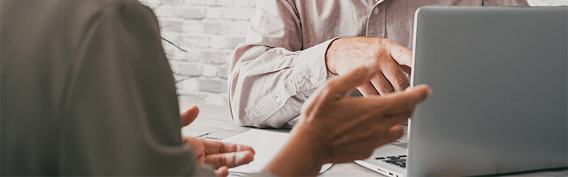 Zwei Personen sitzen an einem Tisch und führen ein Gespräch, während eine von ihnen auf einen Laptop zeigt. Ihre Hände sind im Vordergrund zu sehen.