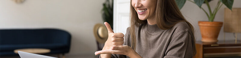 Eine lächelnde Person sitzt in einem hellen Raum und nutzt Gebärdensprache. Im Hintergrund stehen eine Couch, ein kleiner Tisch und eine Pflanze.