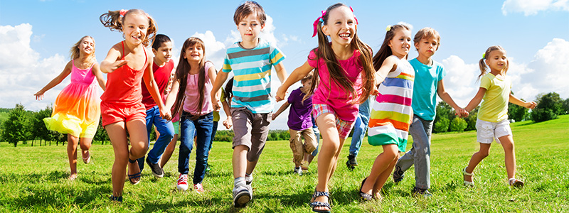 Gruppe fröhlicher Kinder läuft Hand in Hand über eine grüne Wiese bei sonnigem Wetter.